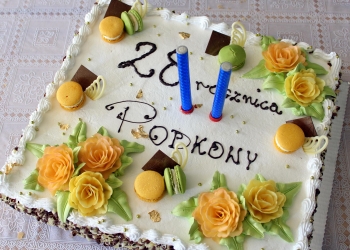 Tort urodzinowy. Na środku świeczki, napis 28 rocznica Podkowy, po bokach ozdoby z cukrowych kwiatów.