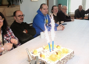 Zapalone świece na torcie urodzinowym. Dookoła tortu przy stole siedzą Uczestnicy spotkania.