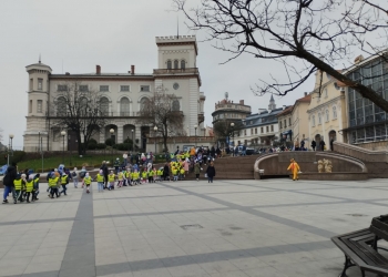 Uczniowie ze szkoły podstawowej, mają na sobie odblaskowe kamizelki, uczestniczą w pochodzie.