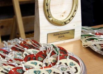 Na stoliku znajdują się pamiątkowe ceramiczne medale oraz złota podkowa.