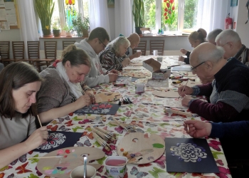 Uczestnicy zajęć malują obvrazki kwiatów i mandali. Stół przy którym pracująjest nakryty obrusem w kolorowe motyle.