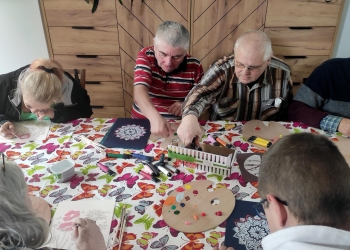 Uczestnicy zajęć malują obvrazki kwiatów i mandali. Stół przy którym pracująjest nakryty obrusem w kolorowe motyle.