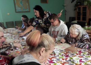 Uczestnicy zajęć malują obvrazki kwiatów i mandali. Stół przy którym pracująjest nakryty obrusem w kolorowe motyle.