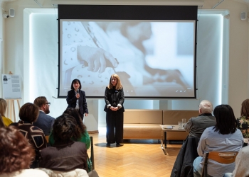 Prelegentka prowadzi wykład, na pierwszym planie osoby siedzą na krzesłach, w tle fotografia dłoni.