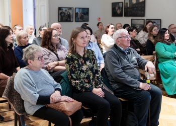 Osoby słuchające wykładu, siedzą na krzesłach, w tle na ścianach wiszą fotografie.