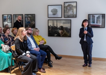 Prelegentka prowadzi wykład, na sali osoby słuchające siedza na krzesłach, w tle na ścianach fotografie.
