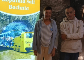 Uczestniczka i uczestnik wycieczki pozują do fotografii stojąc przed tablica informacyjną.