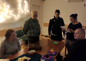 Na fotografii jest stół, wokół którego siedzą i stoja uczestnicy spotkania. jedna z osób odczytuje treść z kartki.