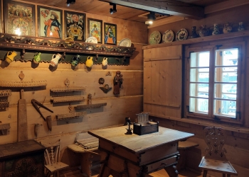 Wnętrze jednej  z izb chałupy Gąsieniców. Na pierwszym planie drewniany stół z ławą, na ścianach święte obrazy i narzędzia drewniane, jest okno z widokiem na podwórze.