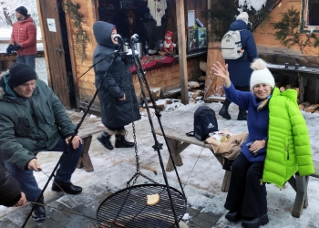 Kilka osób siedzi obok ogniska. Osoby są ciepło ubrane, maja nakrycia głowy i zimowe kurtki.