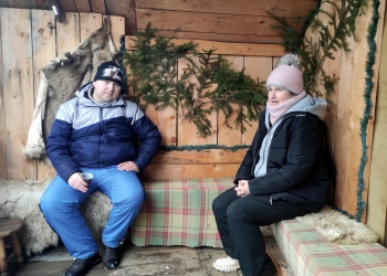 Uczestnicy kuligu siedzą na ławie pod drewnianą ścianą.Na ścianie są zielone gałązki choinki.