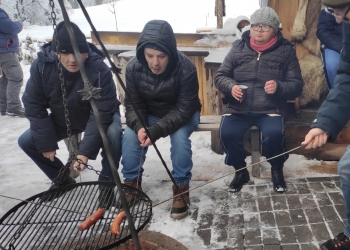Kilka osób siedzi obok ogniska, smażąc kiełbaskę. Osoby są ciepło ubrane, maja nakrycia głowy i zimowe kurtki.