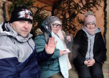 Uczestnicy kuligu siedzą na ławie pod drewnianą ścianą. Na ścianie są zielone gałęzie choinki.