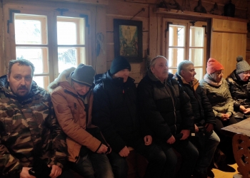 Uczestnicy wycieczki siedzą  na ławie dookoła drewnianego stołu słuchając historii powstania chałupy Gąsieniców.
