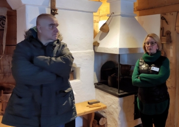 Przewodniczka stojąca obok białego pieca opowiada historię chałupy Gasieniców. Obok niej stoi uczestnik wycieczki.