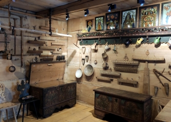 Wnętrze jednej z izb. Znajdują się w niej drewniane sprzęty i skrzynie, na ścianach wiszą święte obrazy i rzeczy potrzebne w domowym gospodarstwie.