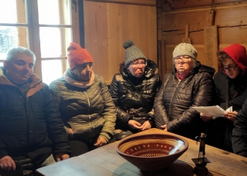 Uczestnicy wycieczki siedzą  na ławie dookoła drewnianego stołu słuchając historii powstania chałupy Gąsieniców. Na stole jest duża gliniana misa, pięknie ozdobiona.