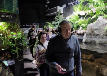 Kilka osób zwiedza Podwodny Świat przechodząc pomiędzy akwariami, Nad akwariami znajdują się zielone rośliny.