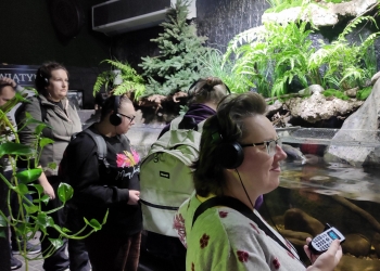 Kilka osób ze słuchawkami na uszach słucha nagrania audio podczas zwiedzania wystawy. W tle akwarium oraz zielone paprocie.