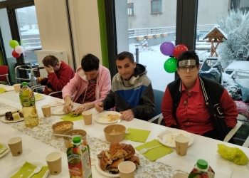 Kilka osób siedzi przy stole. Na stole jest ciasto, napoje i jasno zielone serwetki. W tle okno ozdobione kolorowymi balonami. Za oknem śnieg i karmnik dla ptaków.