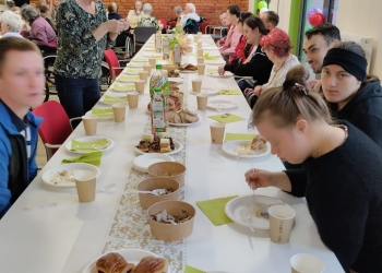 Kilka osób siedzi przy stole. Na stole jest ciasto, napoje i jasno zielone serwetki. 