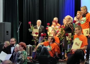 Wspólna fotografia osób wyróżnionych w konkursie. Uczestnicy stoją na scenie. Osoby wyróznione trzymają w rękach dyplomy i czerwone róże.