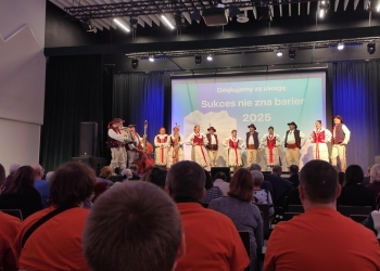 Na pierwszym planie członkowie Klubu Górskiego w ppomarańczowych koszulkach. Na dalszym pplanie scena na której występuje góralski zespół w ludowych strojach.