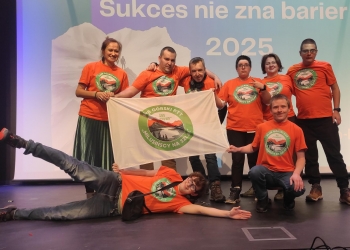 Wspólna fotografia członków Klubu Górskiego. Uczestnicy mają na sobie pomarańczowe koszulki z logo Klubu a dwie osoby trzymają w rękach flagę Klubu. 