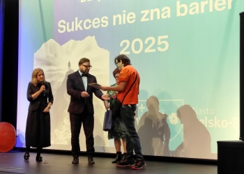 Zastępca prezydenta miasta Bielska-Białej wręcza dyplom wyróżnienia członkowi Klubu Górskiego. Uczestnik ma na sobie pomarańczową koszulkę. w tle baner nawiązujący do treści konkursu.