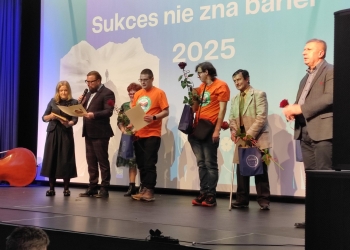 Osoby wyróżnione stoją na scenie, trzymając w rękach dyplomy i czerwone róże.