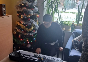 Uczestniczka spotkania gra na przenośnych organach. W tle jest ubrana choinka, oraz duże jasne okno z kwiatami na parapecie.