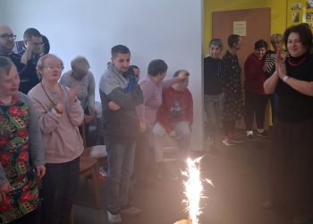 Grupa uczestników spotkania stoi dookoła stołu. Na stole jest tort, w torcie jest płonąca raca tortowa