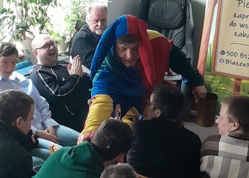 Uczestnicy biora udział w występie Błazna, siedząc na krzesłach. w tle duże, jasne okno.