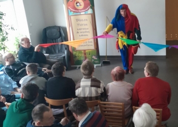 Uczestnicy spotkania siedzą na krzesłach, ogladając występ Błazna.W tle baner nawiazujący do występu, oraz duże jasne okno.