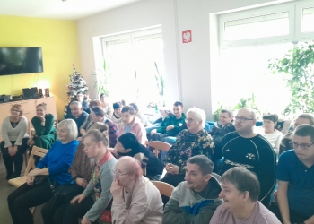 Uczestnicy i ich rodziny siedza na krzesłach W tle są dwa duże, jasne okna. 