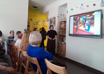 Uczestnicy spotkania siedzą na krzesłach. Pani Dyrektor prowadzi prezentację. W tle na ścianie duży ekran, na którym prezentowane są zdjęcia z wydarzeń z życia filii z ostatniego roku. 