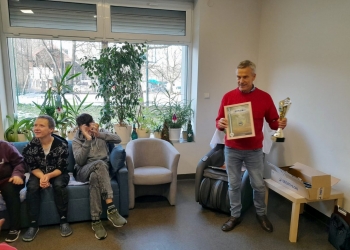 Uczestnicy słuchają przemawiającego prezesa Towarzystwa Miłośników Ziemi Bielsko-Bialskiej. Prezes trzyma w rękach puchar i dyplom, w tle jest duże, jasne okno z kwiatami na parapecie.