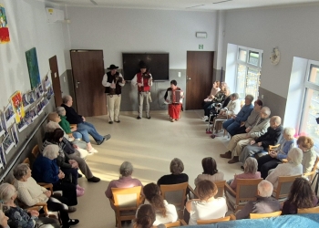 Seniorzy siedzą w kręgu, słuchając kolęd w wykonaniu kapeli. Na dalszym planie członkowie kapeli w strojach ludowych grają na instrumentach.