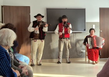 Członkowie kapeli dwóch mężczyzn i kobieta ubrani w stroje ludowe grają na instrumenatach. Mężczyźni grają na skrzypcach a kobieta na akordeonie.