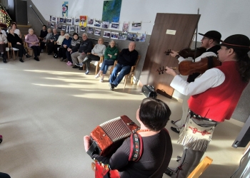 Członkowie kapeli są na pierwszym planie, graja na instrumentach. dookoła siedzą seniorzy śpiewając.