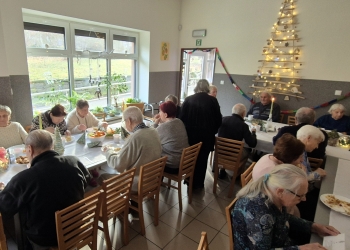 Uczestnicy spotkania siedzą przy stole spożywając posiłek. W tle na scianie jest oswietlona złota choinka.