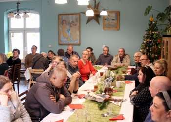 Uczestnicy spotkania siedzą przy stole. Stół jest świątecznie udekorowany, widać świąteczną choinkę.