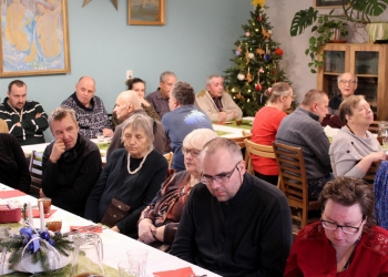 Uczestnicy spotkania siedzą przy stole. Stół jest świątecznie udekorowany, widać świąteczną choinkę.