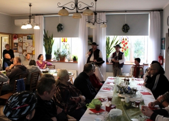 Uczestnicy siedzą dookoła udekorowanego stołu, w tle członkowie Kapeli w strojach ludowych grają na instrumentach.