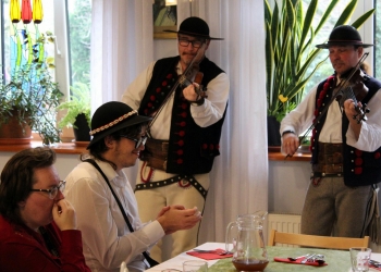 Uczestnicy siedzą przy udekorowanym stole, w tle członkowie Kapeli w strojach ludowych grają na instrumentach.