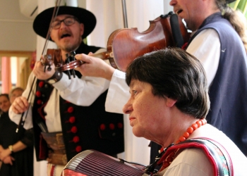 Dwaj mężczyźni w strojach ludowych grają na skrzypcach, kobieta gra na akordeonie, ma na szyi czerwone korale.