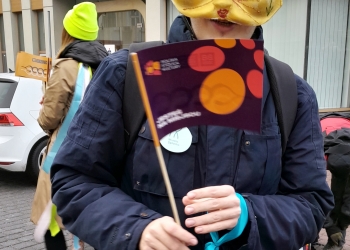 Na zdjęciu jest widoczna osoba w masce kota. trzymająca chorągiewkę.W tle widoczny budynek banku.