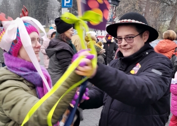 Zdjęcie przedstawia kobietę i mężczyznę biorących udział w pochodzie i tańczących. Kobieta trzyma żółtą wstążkę, mężczyzna ma na głowie góralski kapeusz.