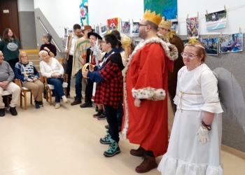 Na pierwszym planie uczestnicy kolędowania ubrani w stroje kolędnicze.Kobieta ma na sobie biały strój i skrzydła anioła, meżczyzna w czerwonym płaszczu ma na głowie koronę, na dalszym planie seniorzy. 