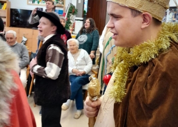 Uczestnik kolędowania ma na ramionach brązowy płaszcz, na głowie koronę. Na dalszym planie osoby ubrane w stroje kolędnicze.